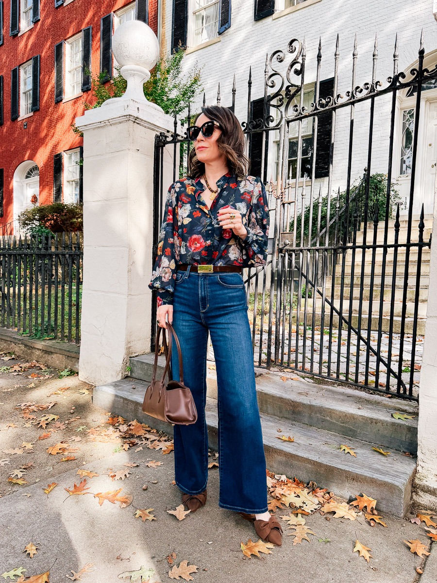October 2026 Outfit Details - dark floral print blouse tucked into high-waisted dark blue wide-leg jeans, styled with a brown belt, two-tone bow-accented flats, a brown shoulder bag, and sunglasses