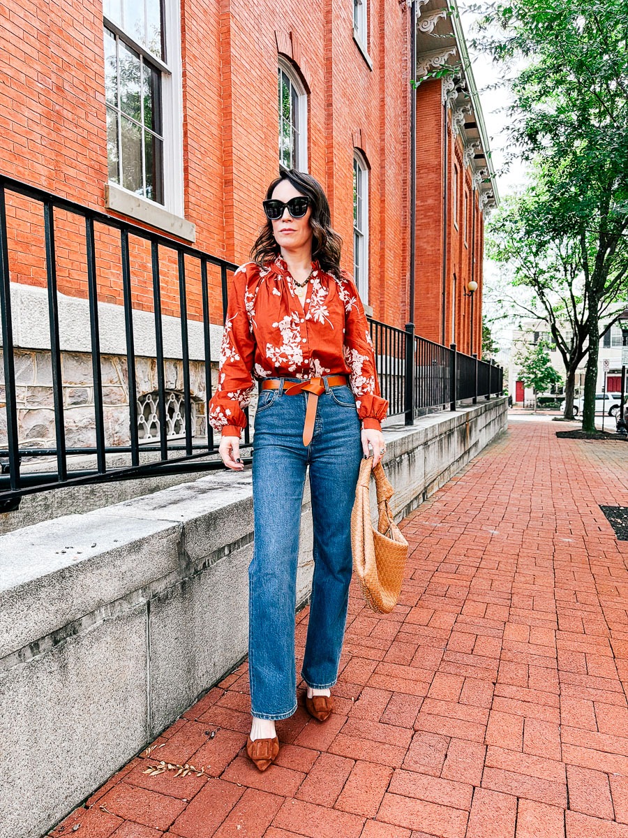 October 2026 Outfit Details - rust-orange floral print blouse paired with straight-leg blue jeans, a tan belt, matching tan pointed-toe flats, and a woven slouchy shoulder bag